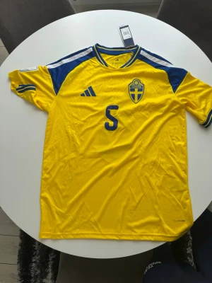 Sverige matchtröja Adidas gul/blå - Säljer en officiell Sverige matchtröja från Adidas i gult med blå detaljer. Tröjan har nummer 5 och namnet Gudmundsson på ryggen, samt landslagets emblem på bröstet. Tillverkad i lätt och ventilerande material, perfekt för fotboll eller träning.