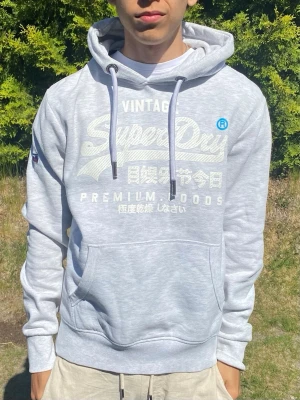 Vit hoodie från Superdry - Ljusgrå hoodie från Superdry med stor vit logga och japansk text på bröstet. Klassisk känguruficka framtill, justerbar huva med vita snören och mjukt, borstat innerfoder. Perfekt för en avslappnad streetwear-look.