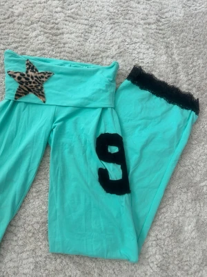 Turkosa loungewear-byxor med spets - Turkosa loungewear-byxor med svart spetskant vid benslutet, leopardmönstrad stjärna på linningen och en svart siffra 9 på ena benet. 