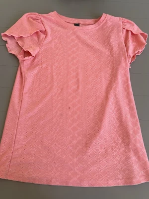 Rosa topp med volangärmar - Söt rosa topp med korta volangärmar och diskret spetsmönster. Toppen har rund halsringning och är tillverkad i ett mjukt material som känns bekvämt mot huden. Perfekt för dig som gillar feminina detaljer och en ljus färgpalett.