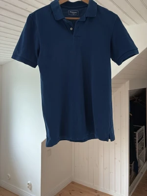Blå pikétröja från Dressmann - Klassisk blå pikétröja från Dressmann med krage och knappar framtill. Tröjan har korta ärmar och är tillverkad i mjuk bomull, perfekt för en avslappnad och stilren look.