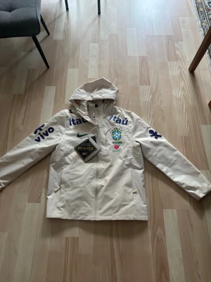 Nike Brasil vindjacka med huva - Snygg vit vindjacka från Nike med Brasil-logga och sponsormärken i blått. Jackan har huva, dragkedja framtill och är tillverkad i GORE-TEX-material som står emot regn och vind. Perfekt för dig som gillar sportig stil och vill sticka ut med landslagskänsla. Är size M men det är närmare size S