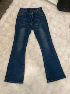Blå bootcut jeans med hög midja - Snygga blå bootcut jeans med hög midja och klassiska fem fickor. Jeansen har en lätt tvättad look och vida ben från knät och ner. Perfekta för dig som gillar retrostil och vill ha ett par jeans som sticker ut lite extra.
