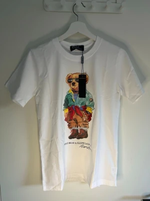Vit Polo Bear t-shirt från Ralph Lauren - Vit t-shirt från Ralph Lauren med ikoniskt Polo Bear-tryck framtill. Björnen har cool outfit med solglasögon och färgglada detaljer. Klassisk rund hals och korta ärmar. Tillverkad i mjuk bomull för skön känsla.