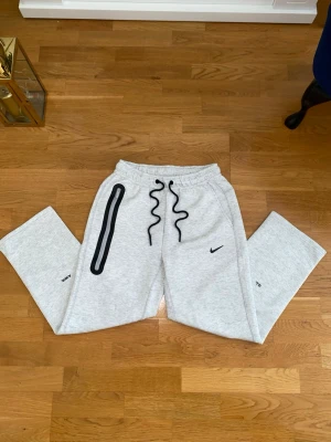 Gråa mjukisbyxor från Nike x AMBUSH - Snygga ljusgrå mjukisbyxor från Nike x Nocta AMBUSH med svart dragsko i midjan och unik svart detalj med dragkedja på ena benet. Byxorna har en relaxed fit och är tillverkade i mjukt sweatshirtmaterial. Perfekta för en chill och trendig look.