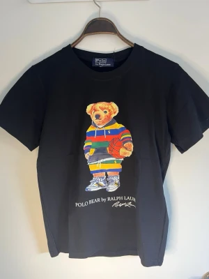 Svart Polo Bear t-shirt från Ralph Lauren - Svart t-shirt från Polo Ralph Lauren med ikoniska Polo Bear-trycket framtill. Björnen har färgglada kläder och håller en basketboll. T-shirten har rund hals och korta ärmar. Klassisk och stilren design med tryckt logga under motivet.