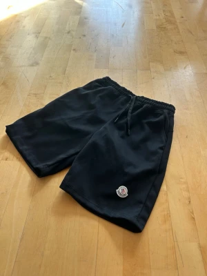 Svarta shorts från Moncler med snörning - Säljer ett par svarta shorts från Moncler med elastisk midja och snörning. Shortsen har en klassisk passform och Moncler-logga på vänstra benet. Perfekta för sommaren och tillverkade i mjuk bomull för maximal komfort.
