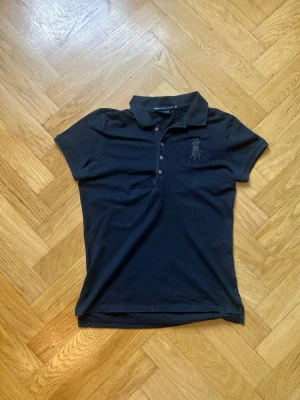 Svart pikétröja från Ralph Lauren - Klassisk svart pikétröja från Ralph Lauren med korta ärmar och knappslå framtill. Tröjan har en broderad logga på bröstet och är tillverkad i mjuk bomull. Perfekt till jeans eller kjol för en sportig och stilren look. Har en liten defekt se på sista bild
