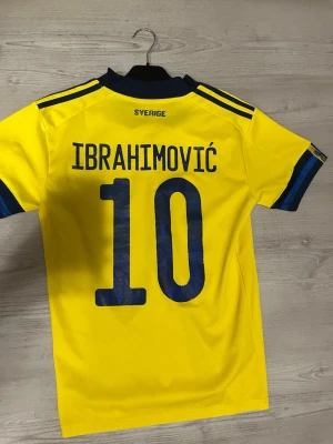 Sverige fotbollströja Ibrahimović 10 - Gul fotbollströja från Adidas med blå detaljer, tryck av 'IBRAHIMOVIĆ' och nummer 10 på ryggen. Tröjan har korta ärmar, blå ränder på axlarna och blå krage. Perfekt för dig som vill stötta svenska landslaget. Men kan passa även för storlek M