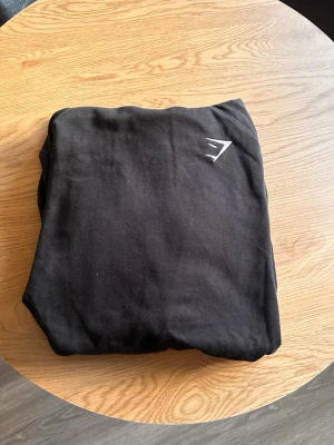 Svart hoodie från Gymshark - Säljer en svart hoodie från Gymshark med deras logga broderad på bröstet. Hoodien har huva med dragsko och en stor magficka. Tillverkad i mjukt bomullsmaterial och har ribbade muddar vid ärmslut och nederkant. Perfekt för chill eller gymmet. Använd ca 3 gånger 