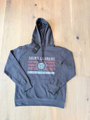 Mörkgrå Saint Laurent hoodie - Snygg mörkgrå hoodie från Saint Laurent med tryck framtill i vitt och rött samt texten 'SAINT LAURENT'. Perfekt för en avslappnad streetwear-look. Tror du vet varför det billiga priset😊