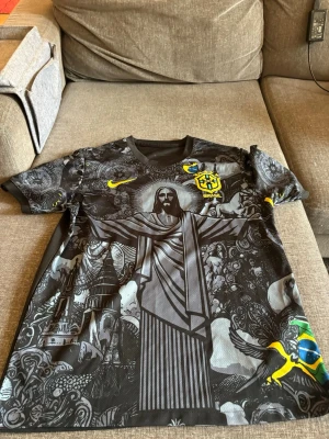 Svart Brasilien fotbollströja Nike - Unik svart Brasilien fotbollströja från Nike med detaljerat tryck av Kristusstatyn och brasilianska symboler. Tröjan har gula Nike-loggan och CBF-emblemet på bröstet. Tillverkad i lätt och ventilerande material, perfekt för träning eller match.