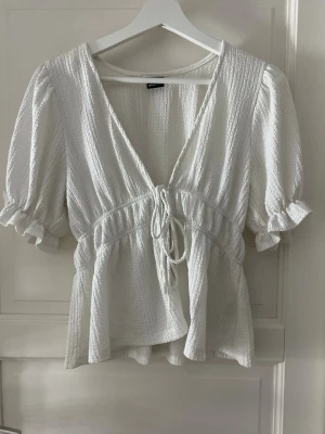 Vit puffärmad blus med knyt - Supersöt vit blus från Gina Tricot med puffiga korta ärmar och volangkant. Blusen har struktur i tyget, v-ringning och knyt framtill i midjan för en feminin siluett. Perfekt till jeans eller kjol för en trendig look.