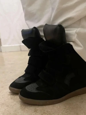 Svarta high top sneakers med kardborre - Säljer ett par svarta high top sneakers med breda kardborreband och tjock plös från Isabel marant. Skorna har en mix av mocka och läderdetaljer samt en brun gummisula. Perfekta för dig som gillar en chunky och sportig look.