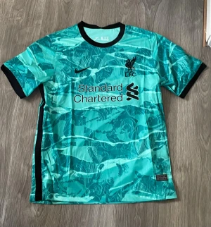 Liverpool FC turkos matchtröja Nike - Snygg turkos matchtröja från Liverpool FC 2020/21 med svart Nike-logga och klubbmärke på bröstet. Tröjan har ett unikt mönster i olika nyanser av turkos, svarta detaljer vid krage och ärmslut samt sponsortryck på framsidan. Perfekt för fotbollsträning eller att bära till vardags.