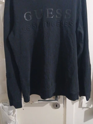 Svart tröja från Guess - Säljer en svart långärmad tröja från Guess med diskret tryck 'GUESS LOS ANGELES' på bröstet. Tröjan har rund halsringning och är tillverkad i mjukt material som känns skönt mot huden. Perfekt för en clean och stilren look.