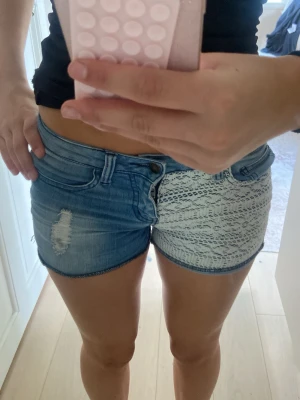 Blå denimshorts med spetsdetalj - Unika blå denimshorts med ena sidan i klassisk jeans och andra sidan täckt av vit spets med mönster. Shorts med låg midja, slitning på jeansdelen och snygg kontrast mellan materialen. Perfekta för sommarens alla äventyr!