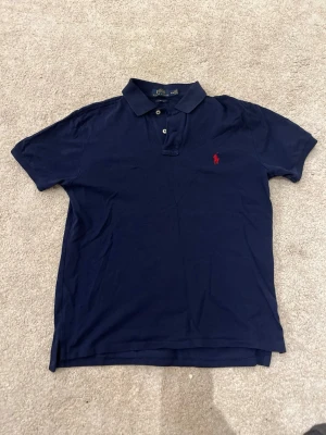 Marinblå pikétröja från Polo Ralph Lauren - Klassisk marinblå pikétröja från Polo Ralph Lauren med korta ärmar och krage. Tröjan har två knappar framtill och den ikoniska röda logotypen broderad på bröstet. Tillverkad i mjuk bomull för en bekväm känsla.