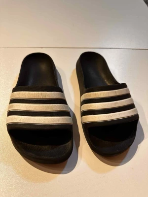 Svarta Adidas badtofflor strl 31 med ränder tofflor  - Klassiska svarta badtofflor från Adidas med tre vita ränder över foten.   Se mått på bilderna  De vita har små "sprickor" som synes på bilderna   Innetofflor Inneskor  Badtofflor Barn Skor