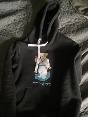 Svart hoodie Polo Bear Ralph Lauren - Svart hoodie från Ralph Lauren med den ikoniska Polo Bear-trycket framtill. Björnen har på sig vit tennisklädsel och håller en dryck och tennisracket. Hoodien har vit dragsko i huvan och en klassisk känguruficka. Perfekt för en sportig och avslappnad stil.