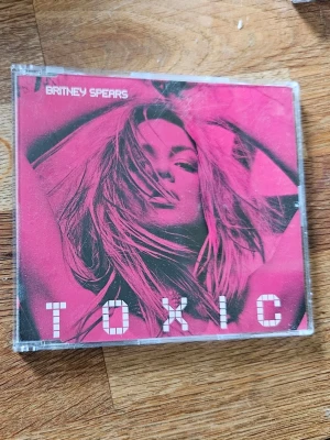Britney Spears Toxic CD-singel - CD-singel med låten 'Toxic' av Britney Spears. Omslaget är rosa med grafiskt tryck och artistens namn i vitt. Skivan ligger i ett genomskinligt plastfodral. Perfekt för dig som gillar popmusik och vill ha en ikonisk låt i samlingen.