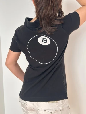 Svart pikétröja från Tommy Hilfiger - Svart pikétröja med klassisk krage och knappar framtill. På ryggen finns en stor vit 8-ball design. Tröjan har korta ärmar och är gjord i mjuk bomull.