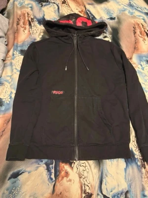 Svart hoodie med dragkedja från Hugo - Säljer en svart hoodie från Hugo med dragkedja och huva. Stort rött Hugo-tryck på huvan och mindre logga på bröstet. Praktiska fickor framtill och justerbar dragsko i huvan. Perfekt för en cool och avslappnad stil.