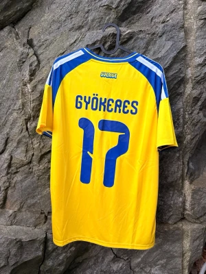 Sverige matchtröja Gyökeres 17 - Gul och blå Sverige matchtröja  med Gyökeres och nummer 17 på ryggen. Tröjan har klassiska blå ränder på axlarna, Tillverkad i lätt och ventilerande material, perfekt för fotboll eller supporterhäng.