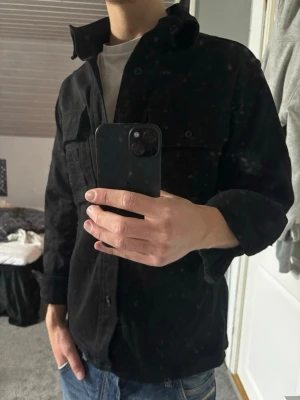 Overshirt  - Säljer en svart overshirt med klassisk krage och två bröstfickor med lock. Overshirten har knäppning framtill och är gjord i ett mjukt, lite tjockare tyg som passar perfekt till lager på lager. Enkel och stilren design som funkar till det mesta.