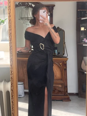 Svart offshoulder långklänning - Elegant svart långklänning med offshoulder-design och v-ringning Klänningen har en markerad midja med dekorativt spänne och en slits framtill som ger en snygg detalj. Köpt från ett märke som heter nelbae. Nypris på denna är 499. Endast testad.