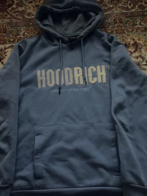 Blå hoodie från Hoodrich med tryck - Blå hoodie från Hoodrich med stor vit logga och texten 'PRODUCT OF THE STREETS' på bröstet. Klassisk känguruficka framtill och justerbar huva med snörning. Mjuk insida och relaxed fit, perfekt för chill dagar.