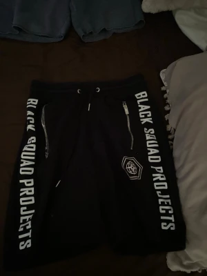 Svarta shorts Black Squad Projects - Svarta shorts från Black Squad Projects med vita tryckta loggor längs båda benen. Shortsen har snörning i midjan och dragkedjor på fickorna. Perfekta för en avslappnad streetwear-look.