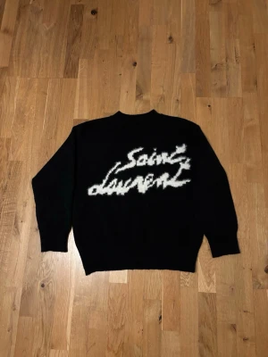Svart Saint Laurent stickad tröja - Svart stickad tröja från Saint Laurent med vit, fuzzy logga i graffiti-stil över bröstet. Tröjan har rund halsringning och långa ärmar. Perfekt för dig som vill ha en statement piece i garderoben.