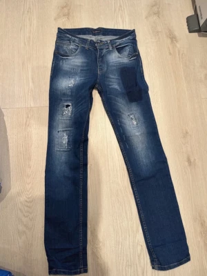 Skinny Jeans Dsquared2 - Säljer ett par blå skinny jeans med slitningar och patchdetaljer på ena benet. Jeansen har klassisk femficksmodell, låg midja och är tillverkade i stretchigt denim. Perfekta för en avslappnad och trendig look. Sparsamt använda. W32 L34, är 183 cm lång och har S/M. 