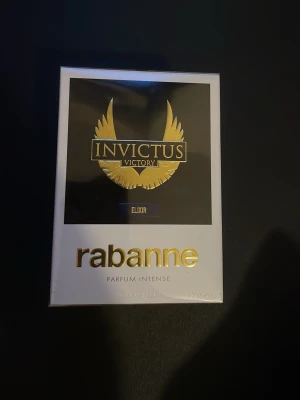 Rabanne Invictus Victory Elixir Parfym - Rabanne Invictus Victory Elixir Parfum Intense är en lyxig herrdoft i elegant svart och vit förpackning med guldiga detaljer. Flaskan har en modern och stilren design, och parfymen är känd för sin intensiva och maskulina doftprofil.