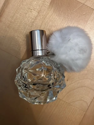 Ariana Grande parfym ”Ari” - Säljer en Ariana Grande parfym i en kristallformad, transparent glasflaska med silverfärgat lock och en stor vit fluffig boll som dekoration. Flaskan har en modern och lyxig känsla, perfekt för dig som gillar snygga detaljer på parfymhyllan.
