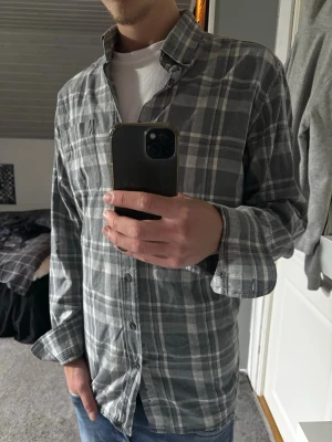 Grå rutig skjorta - Säljer en grå och vit rutig skjorta i mjuk bomull. Skjortan har klassisk krage, knäppning framtill och långa ärmar. Perfekt för en avslappnad och stilren look.