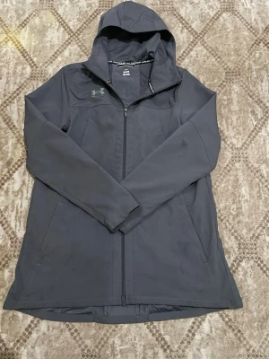 Mörk grå vindjacka från Under Armour - Snygg mörk grå vindjacka från Under Armour med huva och dragkedja framtill. Jackan har två sidofickor och logga på bröstet. Perfekt för blåsiga dagar och sportiga outfits. Materialet är lätt och vattenavvisande.