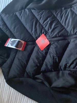 Svart dunjacka från Swedemount - Snygg svart dunjacka från Swedemount med chevronmönstrad vaddering och dragkedja framtill. Jackan har huva och släta ärmar i softshell-material för extra komfort. Perfekt för kalla vinterdagar.