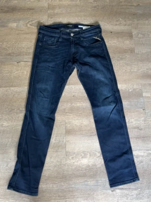 Replay ambass - Snygga mörkblå slim fit jeans från Replay med klassisk femficksdesign och diskreta slitningar. Jeansen har raka ben och tydliga kontrastsömmar. Märkeslapp bak i midjan och dragkedja i gylfen. Perfekta för en trendig och avslappnad look.