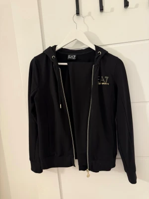 Svart hoodie från EA7 Emporio Armani - Svart hoodie från EA7 Emporio Armani med dragkedja, huva med snören och guldfärgade detaljer. Logga på bröstet och liten guldfärgad logga bak. Tillverkad i mjukt bomullsmaterial och har ribbade muddar vid ärmslut och nederkant. Ett par byxor följer med, det hör ihop 
