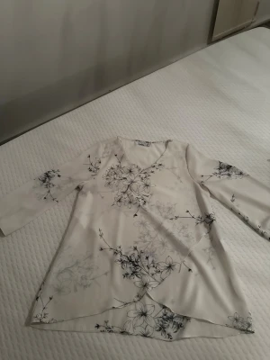 Vit blommig blus från Isolde - Snygg vit blus från Isolde med grå blommönster och v-ringning. Blusen har trekvartsärm och är tillverkad i ett lätt, mjukt tyg som faller fint. Perfekt för dig som gillar en stilren och feminin look.