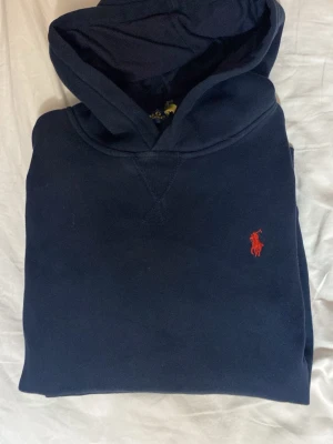 Mörkblå hoodie från Polo Ralph Lauren - Snygg mörkblå hoodie från Polo Ralph Lauren med klassisk röd logga broderad på bröstet. Hoodien har en stor huva och är gjord i mjukt bomullsmaterial. Perfekt för en avslappnad och stilren look.