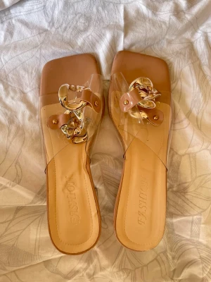 Transparenta sandaler med klack och kedja - Säljer ett par trendiga sandaler från FASHION med transparent rem och chunky guldfärgad kedjedetalj. Sandalerna har fyrkantig tå och genomskinlig blockklack för en modern look. Perfekta för dig som vill sticka ut med stilrena detaljer.