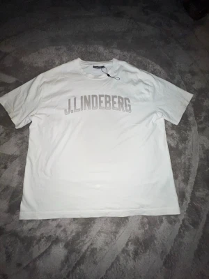 Vit t-shirt från J.Lindeberg - Vit t-shirt från J.Lindeberg med tryckt logga över bröstet. Klassisk passform med rund halsringning och korta ärmar. Perfekt för en stilren och enkel look.