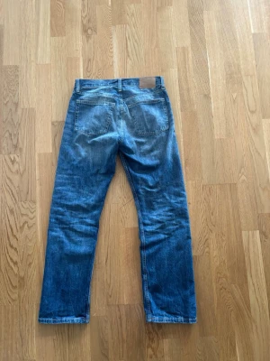 Blå raka denim vintage jeans från Weekday - Säljer ett par klassiska blå jeans från Weekday med raka ben och normal midja. Jeansen har två bakfickor och snygga slitningar för en avslappnad look. Tillverkade i denim med tydliga sömmar och läderpatch med logga bak.