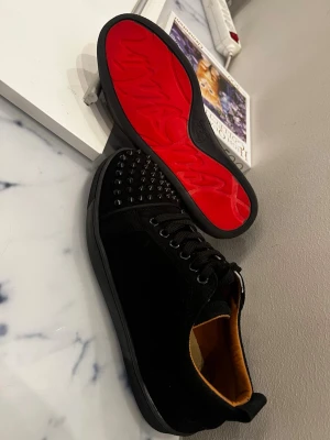 Svarta Louboutin sneakers med nitar - Säljer ett par svarta sneakers från Christian Louboutin med klassisk röd sula och svarta nitar på tån. Skorna har snörning, låg profil och är tillverkade i mocka och läder. Perfekta för dig som vill sticka ut med en edgy och lyxig stil.