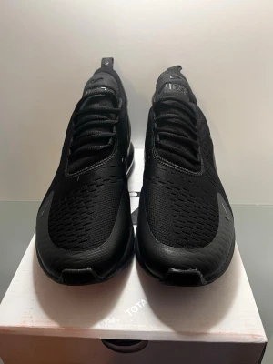 Nike Air Max 270 svart sneaker - Säljer ett par helsvarta Nike Air Max 270 sneakers med mesh-överdel och synlig Air Max-dämpning i hälen. Skorna har en sportig design med snörning och diskreta loggor på tungan och sidan. Perfekta för dig som gillar stilrena och bekväma sneakers.