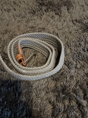 Grått flätat bälte med metallspänne - Snyggt grått flätat bälte med ett stilrent metallspänne och en liten detalj i brunt. Perfekt för att ge din outfit en extra touch. Bältet är flexibelt och passar till många olika stilar.