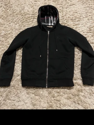 Svart hoodie från Burberry - Svart hoodie med dragkedja från Burberry. Klassisk modell med huva och dragsnören, samt Burberrys rutiga mönster på insidan av huvan. Två fickor framtill och ribbade muddar. Perfekt för en stilren och avslappnad look.